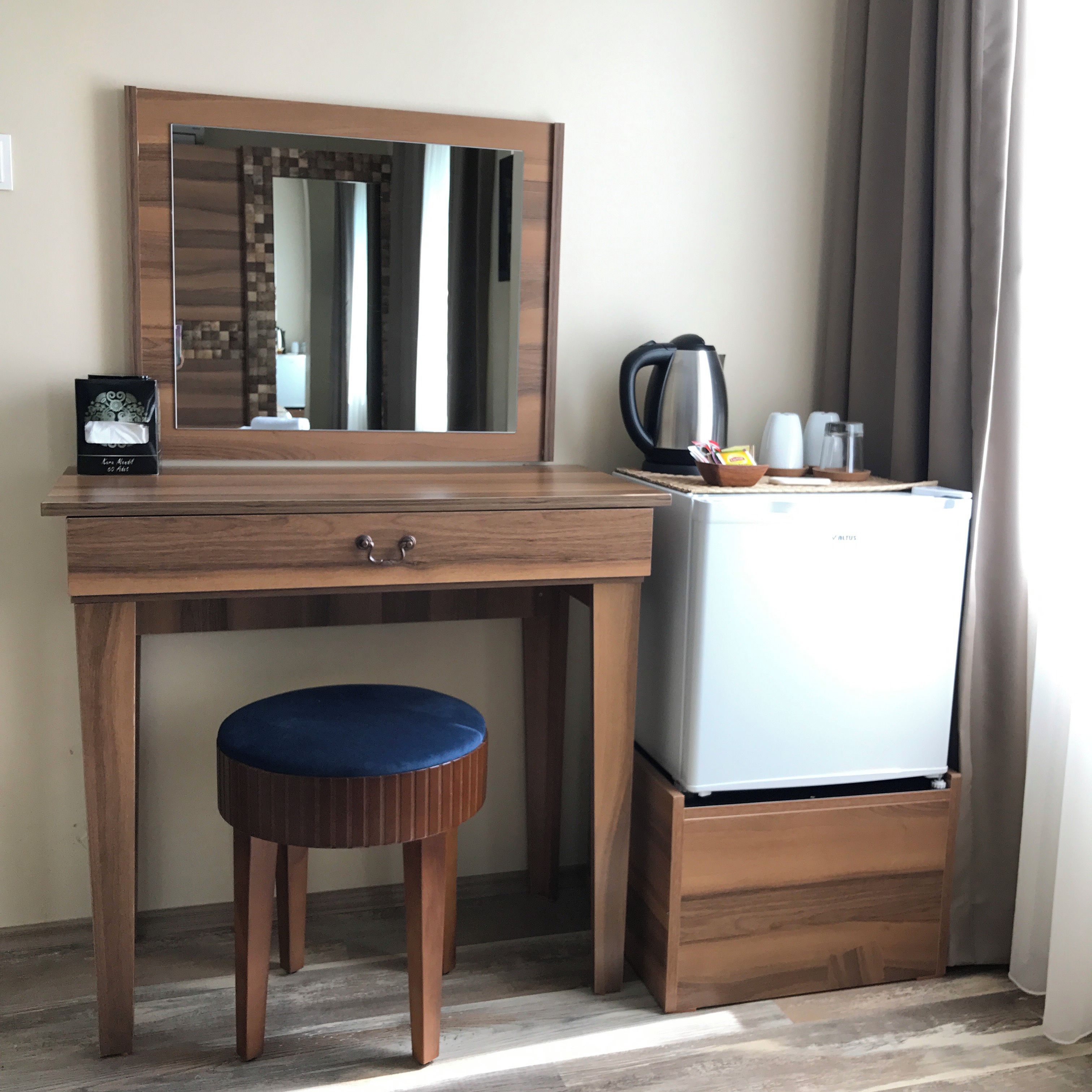A compact room with a wooden desk, a stool, a small refrigerator, a kettle, and a mirror.