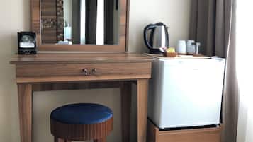 A compact room with a wooden desk, a stool, a small refrigerator, a kettle, and a mirror.