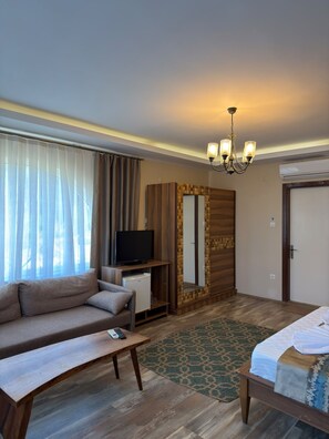 A hotel room with a sofa, a television, a small table, a bed, a mirror, and a chandelier.