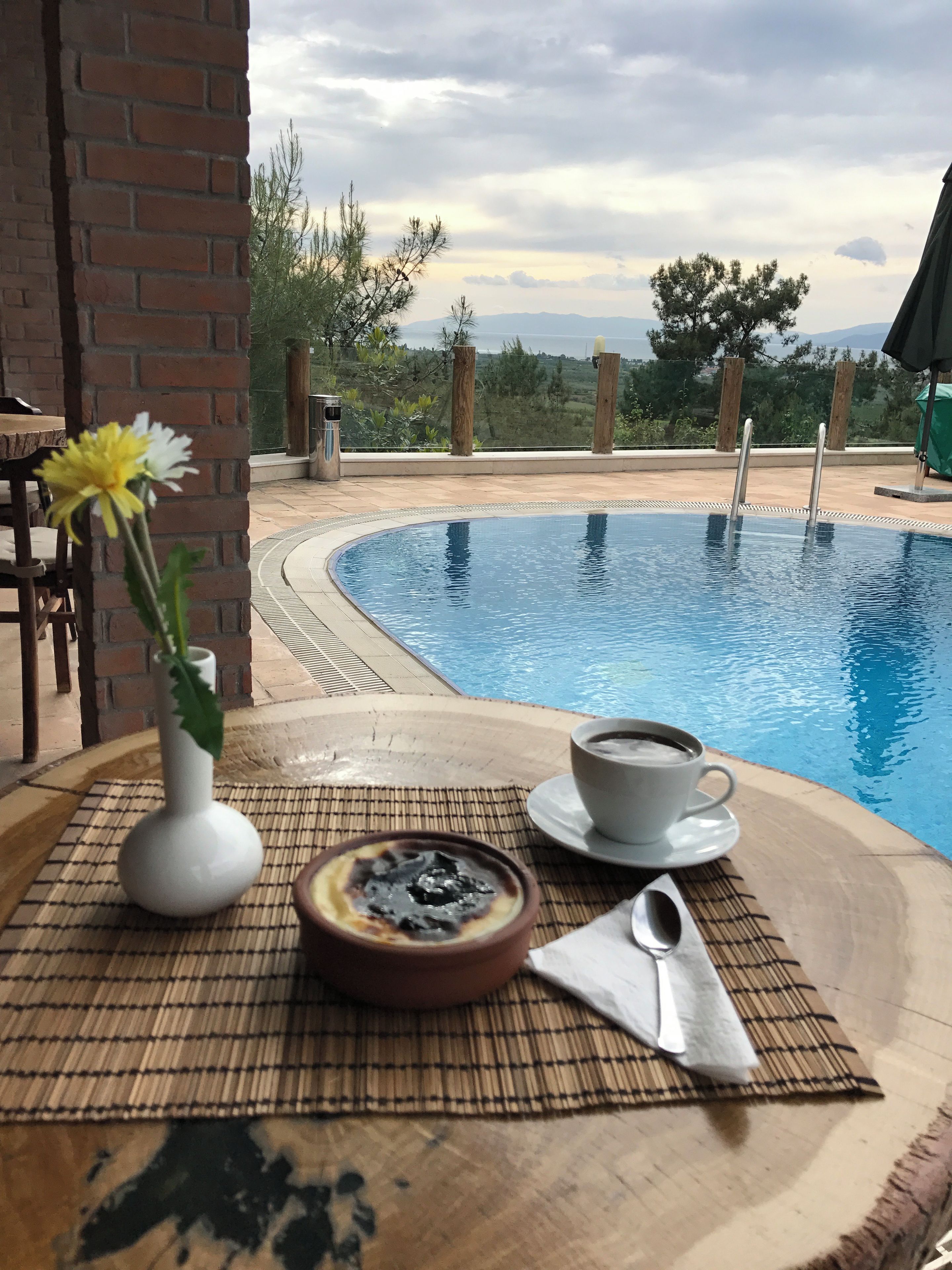 A poolside setting with a table, a white vase with a flower, a cup of coffee, and a dessert bowl.