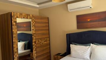 A hotel room with a bed, a wardrobe, a bedside table with towels, an air conditioning unit, and a wall-mounted artwork.