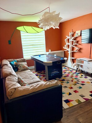 Ein Zimmer mit Billardtisch, einer Couch mit Kissen, einem farbenfrohen Teppich, einer Holz-Baumdekoration und einem Fernseher an der Wand.