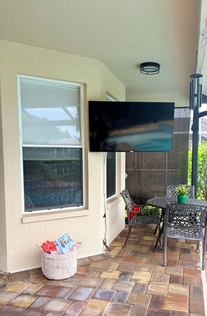 Ein überdachter Patio-Bereich mit an der Wand montiertem Fernseher, einem Korbwäschesessel und einem Tisch mit Stühlen.