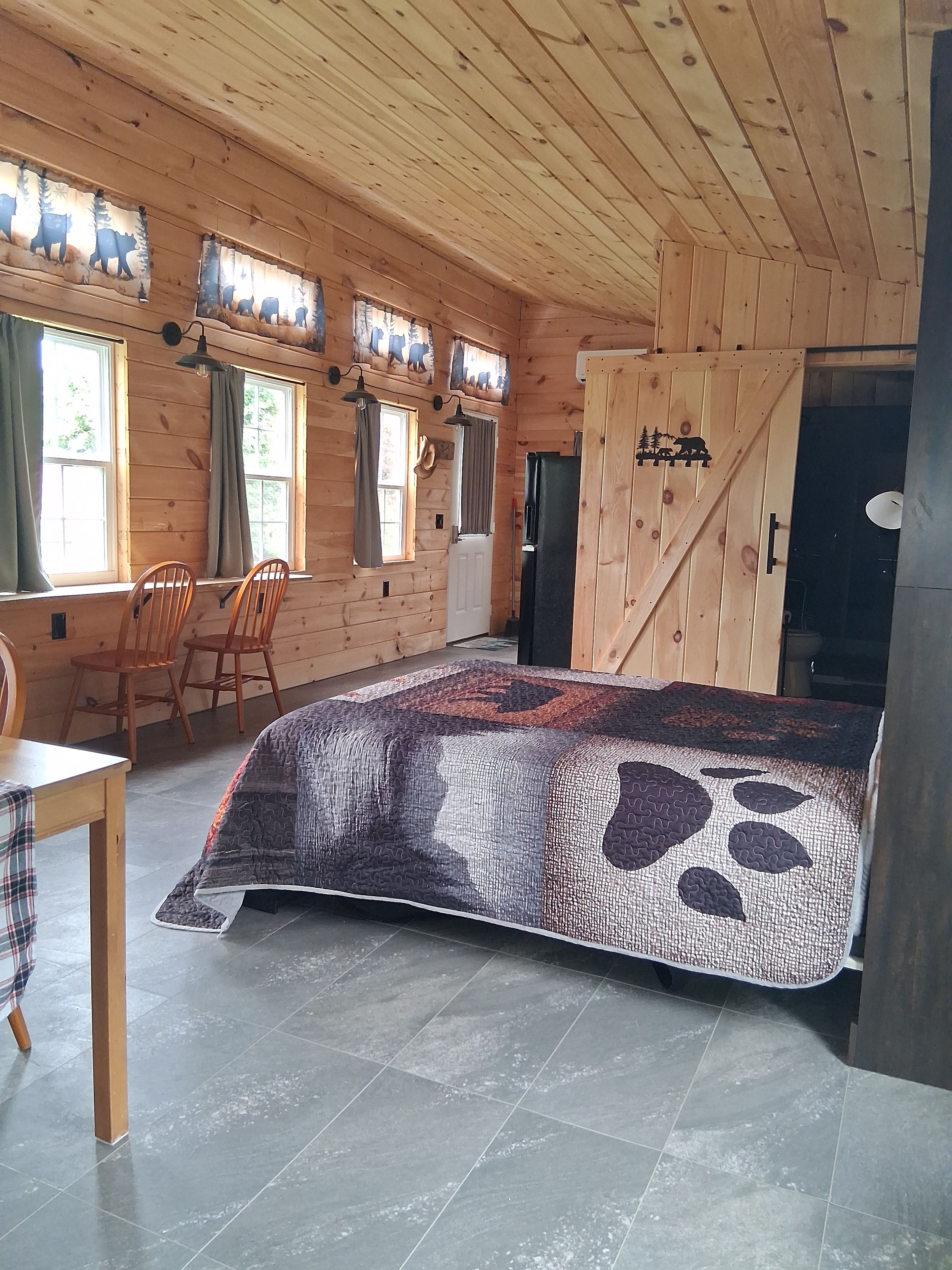 A wooden cabin interior with a bed, two chairs, a table, and a door.