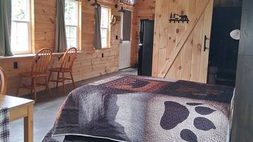 A wooden cabin interior with a bed, two chairs, a table, and a door.