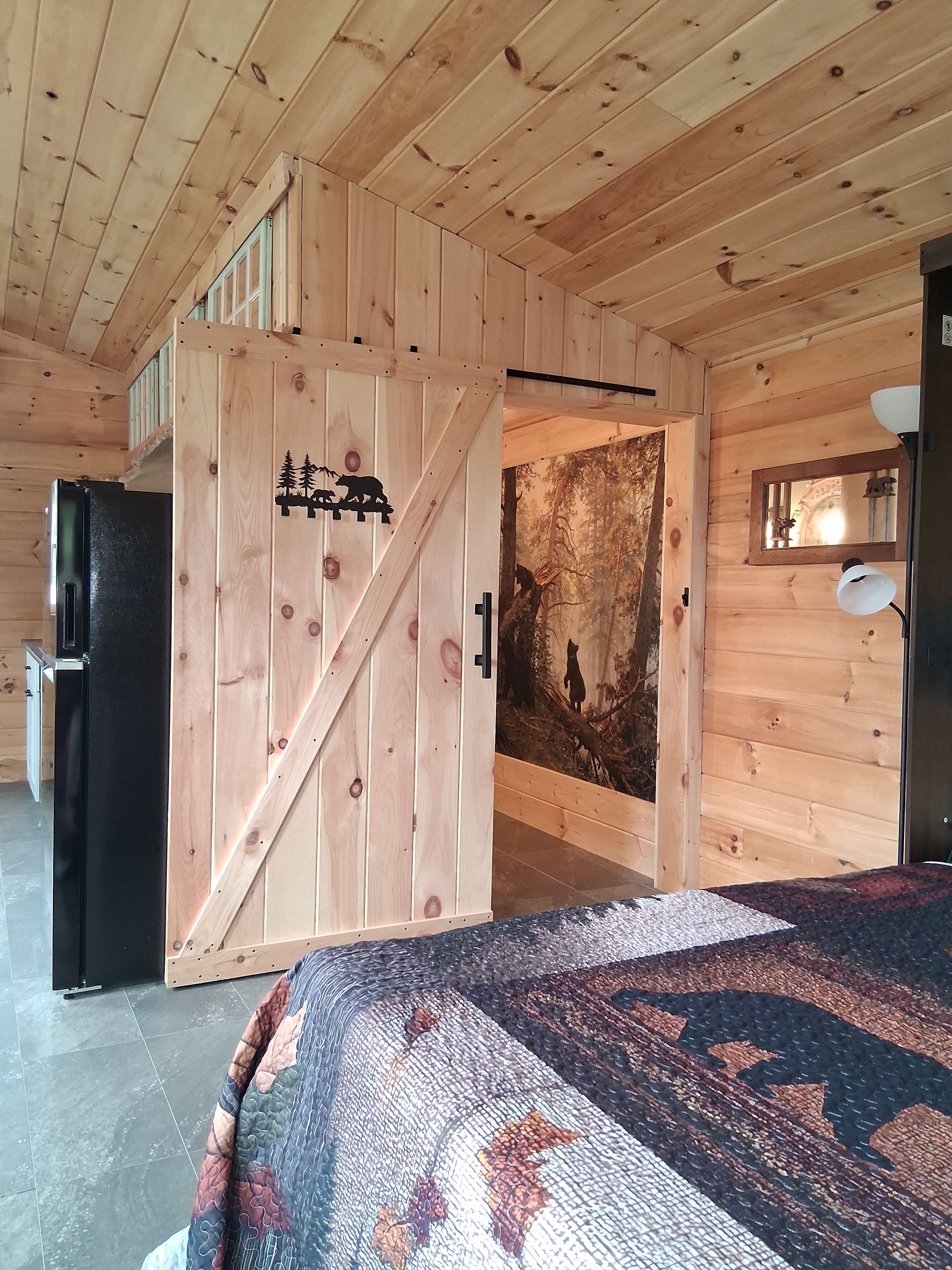 A wooden interior with a bed, a refrigerator, and a door with a forest mural.