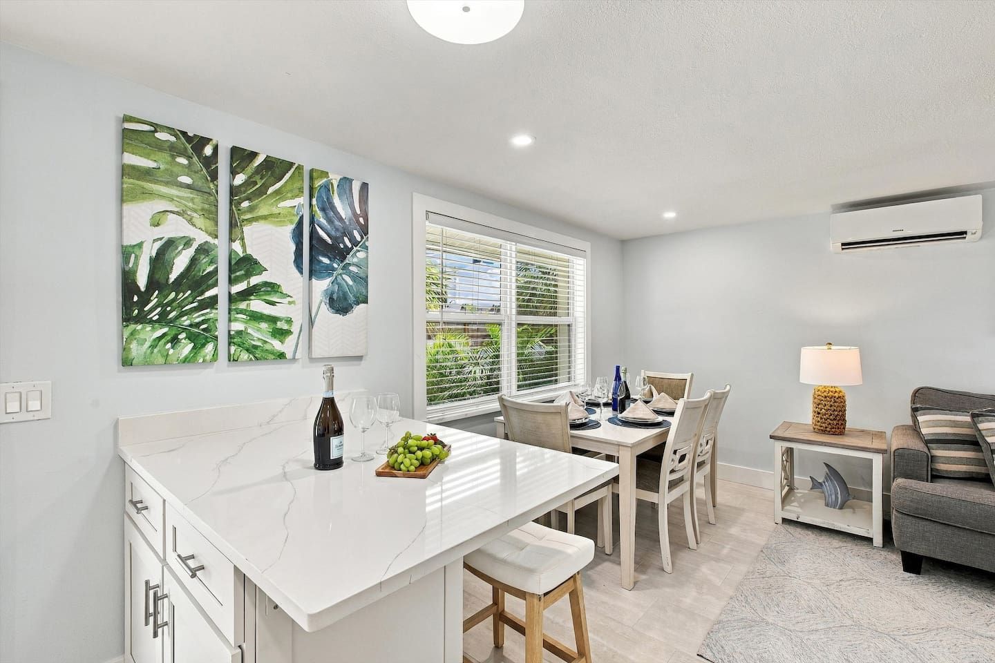 A modern kitchen with a marble countertop, dining area with chairs, a table set for two, and a grey sofa in the living area.