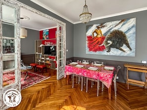 A dining area with a long table set for six, a red patterned tablecloth, and a large painting of a woman and a bear on the wall. - Cozy gîte in La Chaume - 6 guests (Les Sables-d'Olonne)
