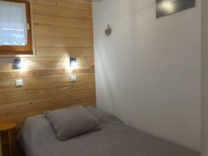 Een kleine kamer met een enkel bed, een houten wand, een raam met gordijn en een wandlamp.