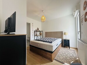 A modern bedroom with a bed, a television on a stand, a unique pendant light, and a radiator. - Air-conditioned apartment with terrace - 2 guests (Balaruc-les-Bains)