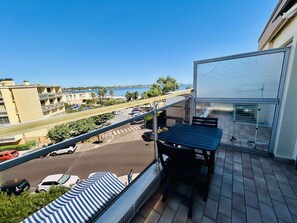Ein Balkon mit Blick auf eine Straße und ein Gewässer. Auf dem Balkon befinden sich ein Tisch und ein Stuhl.