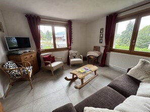 A living room with a television, two armchairs, a sofa, a wooden coffee table, and a view of greenery outside. - Apartment in chalet - 6 guests (Les Carroz d'Arâches)