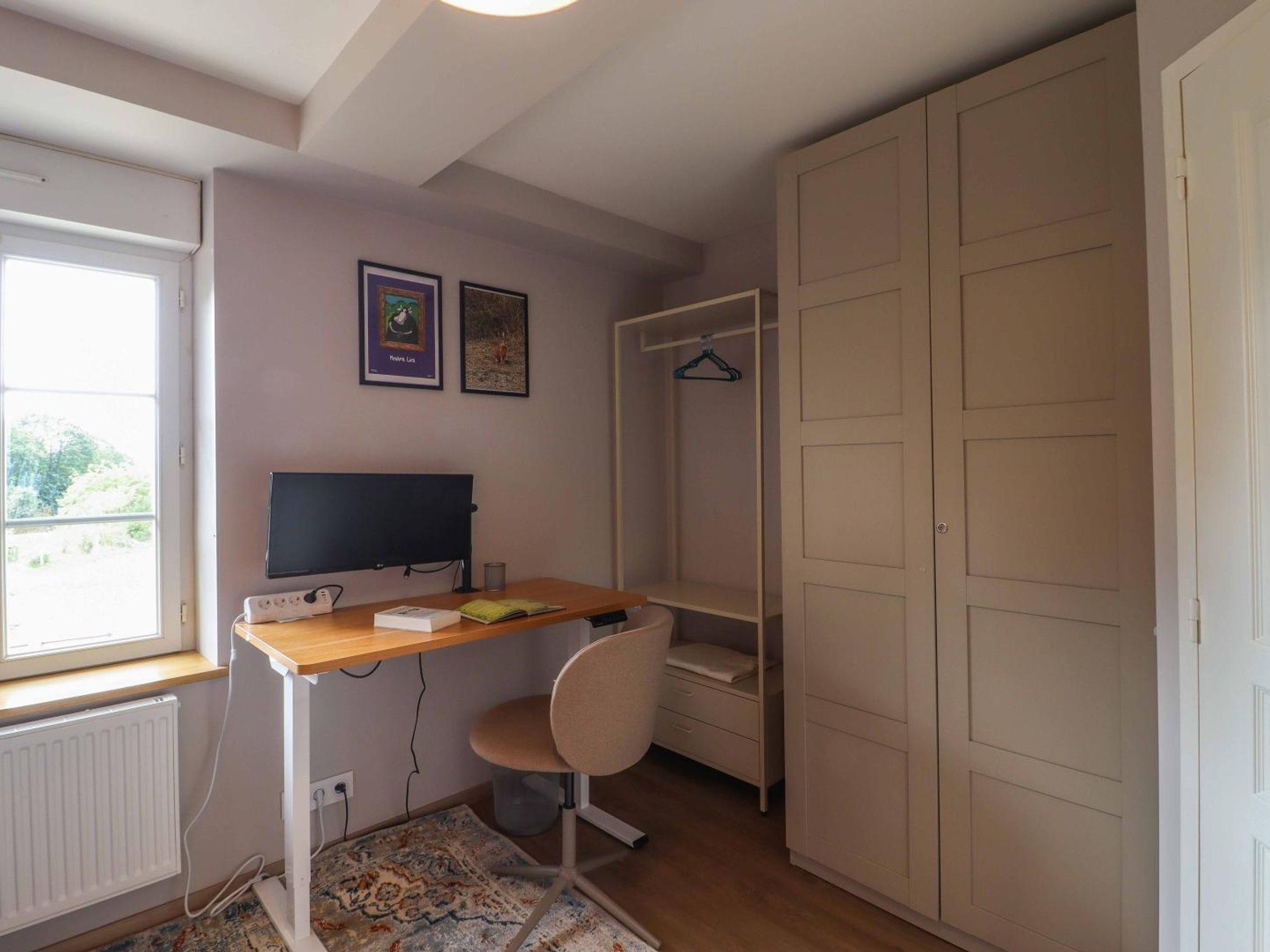 A bedroom with a bed, a desk with a computer, a chair, a wardrobe, and a window.