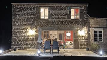 Un bâtiment en pierre doté d’un patio comprenant deux tables et des chaises, un espace de repos couvert et un intérieur visible avec une porte rouge.