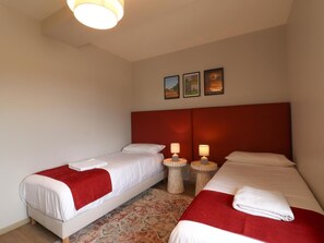 Une chambre d’hôtel avec deux lits simples, chacun recouvert d’une couverture rouge et agrémenté d’oreillers blancs, une petite table ronde avec une lampe et un luminaire au plafond.