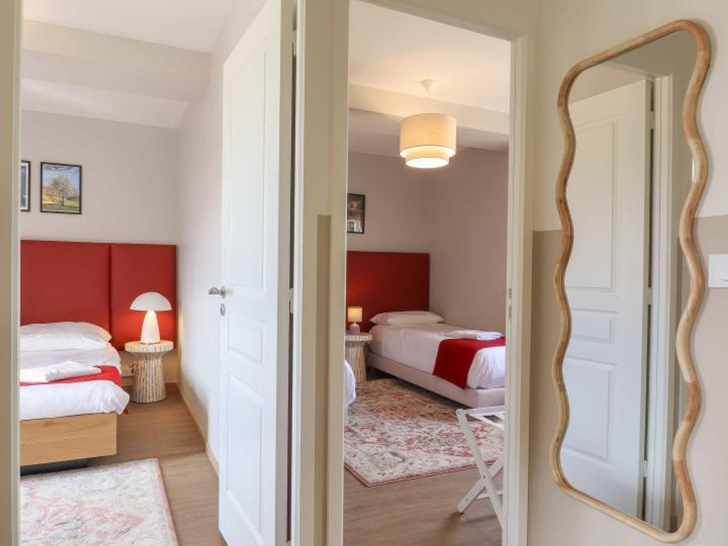 A hotel room with two beds, a red headboard, a bedside lamp, a mirror, and a hanging light fixture.