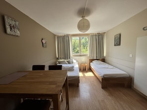 A room with a wooden table, two single beds, a small bedside table, and a window with curtains. - Charming studio with parking - 3 guests, near slopes (Le Lioran)