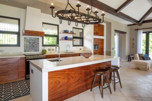 A modern kitchen with a large island, wooden cabinetry, and a hanging light fixture. - Exquisite Spanish Colonial Beachfront Villa: lush gardens    & swimming pool (Savegre)