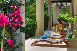 A covered outdoor seating area with lounge chairs and a table, surrounded by lush greenery and flowering plants. - Beautiful beachfront villa with Spanish architecture (Savegre)