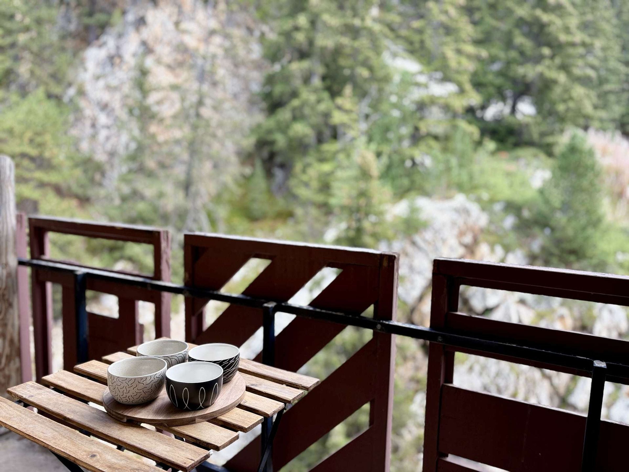Une table en bois, sur laquelle sont posés des bols, se trouve sur un balcon donnant sur une zone boisée.