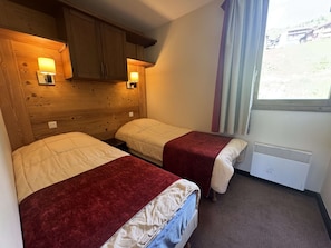 A room with two beds, wooden paneling, a window with curtains, and a wall-mounted heater. - Apartment mountain view in Belle Plagne - 4 guests (Belle Plagne)