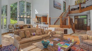 A spacious living room with a large brown sofa, a coffee table with board games, and a view of a kitchen and dining area through large windows. - Mountain Retreat + Pet Friendly + Game Room + Community Hot Tub | Wild Iris Lodg (Edwards)