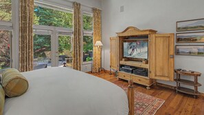 Ein Schlafzimmer mit einem großen Bett, einem Fernseher auf einer Holzkommode und Blick ins Freie durch das Fenster.