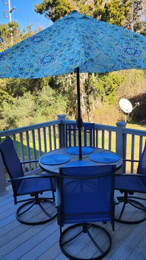Terraza con una mesa y sillas bajo un paraguas estampado, rodeada por una baranda y follaje.