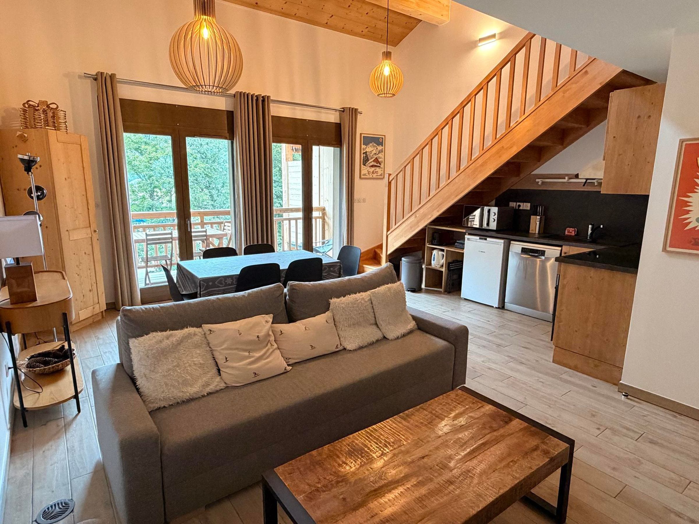 Un salon confortable avec un canapé, une table basse et une table à manger. On aperçoit, au fond, une cuisine avec un escalier en bois.