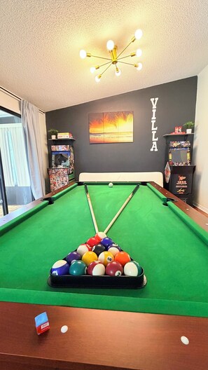 Ein Billardtisch mit grüner Spielvliesfläche, umgeben von einer dunklen Wand mit einem 'VILLA'-Schild, einer Deckenleuchte und einem Fenster mit Vorhängen.