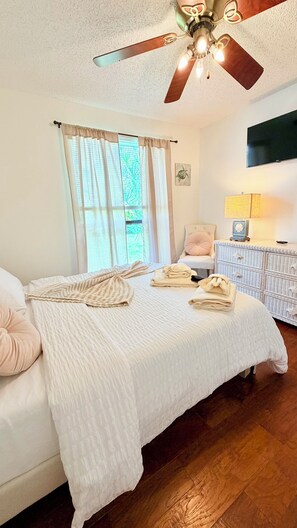 Une chambre à coucher avec un lit, un ventilateur de plafond, une fenêtre avec des rideaux, une table de chevet avec une lampe et un téléviseur fixé au mur.