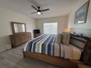 Ein Schlafzimmer mit Bett, Kommode, Fernseher, Deckenventilator und einem Fenster mit Jalousien.