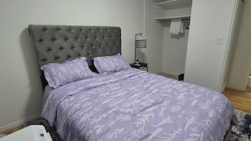 A bedroom with a tufted headboard, a bed with a floral-patterned bedspread, a bedside table with a lamp, and a heater on the floor.