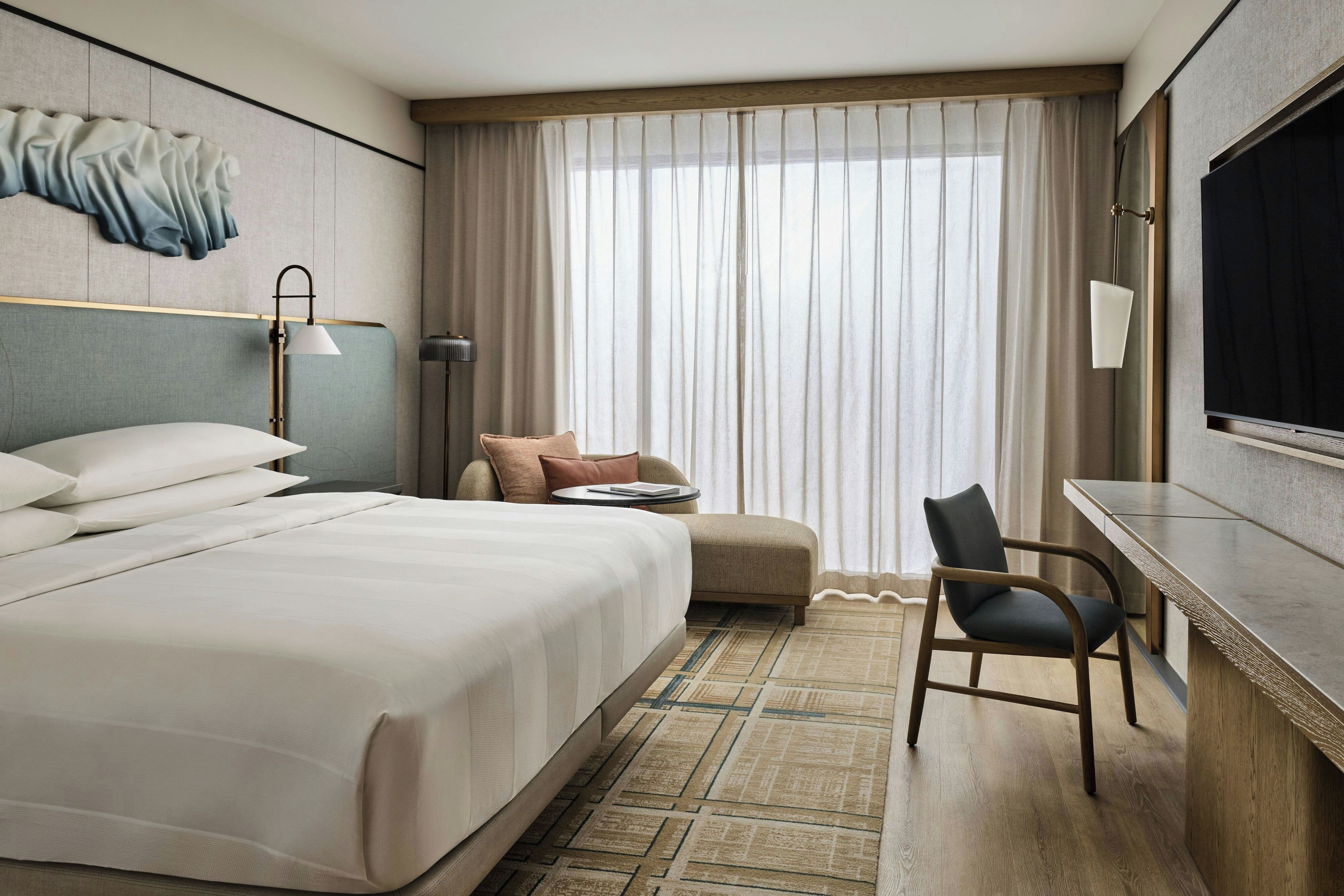 Habitación de hotel con una cama grande, un escritorio, una silla, un televisor y una ventana con cortinas.