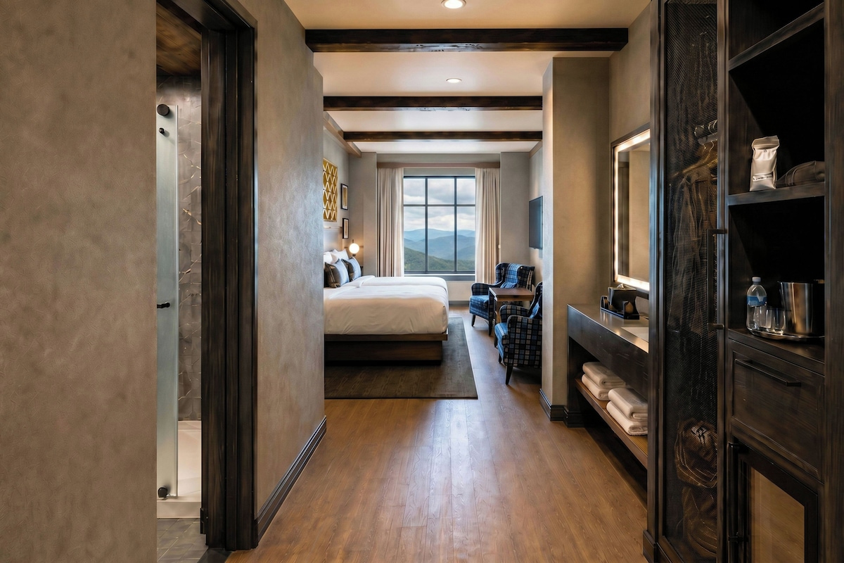 A hotel room with a large bed, a desk with a chair, a large window with a mountain view, and a bathroom with a shower and toilet.