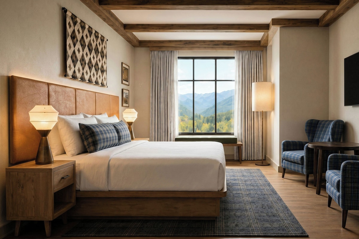 A hotel room with a large bed, two bedside tables with lamps, a seating area with chairs, a view of mountains through the window, and a mounted artwork on the wall.