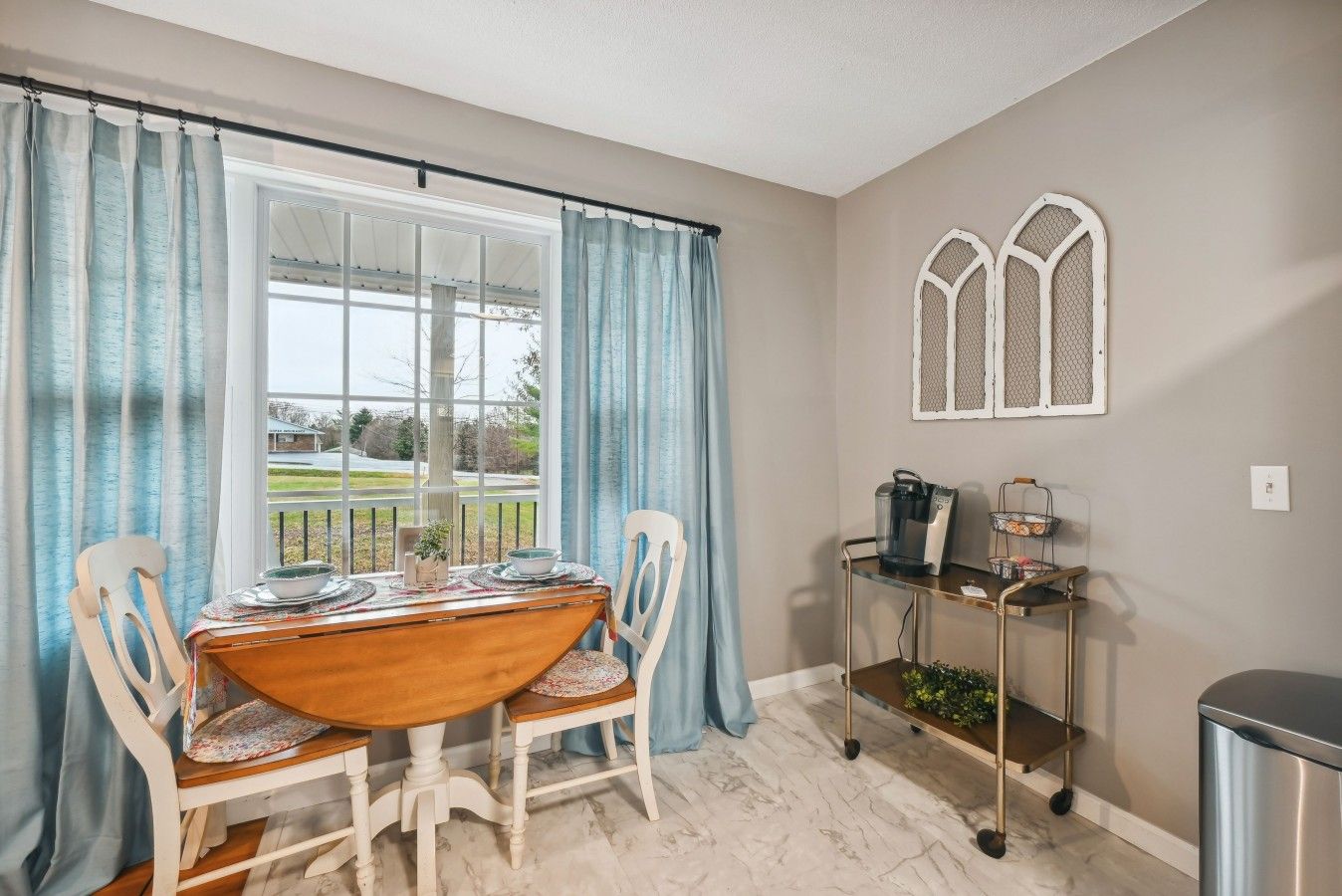 A dining area with a table, chairs, a window with curtains, a coffee station, and a wall-mounted window grille.