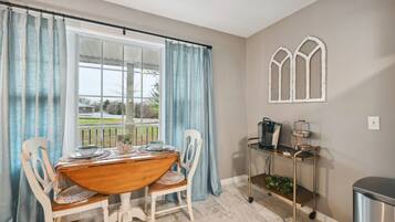A dining area with a table, chairs, a window with curtains, a coffee station, and a wall-mounted window grille.
