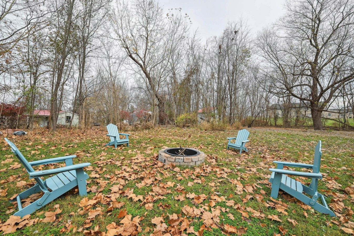 A backyard with three Adirondack chairs, a fire pit, and a grassy area with fallen leaves.