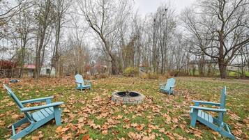 A backyard with three Adirondack chairs, a fire pit, and a grassy area with fallen leaves.