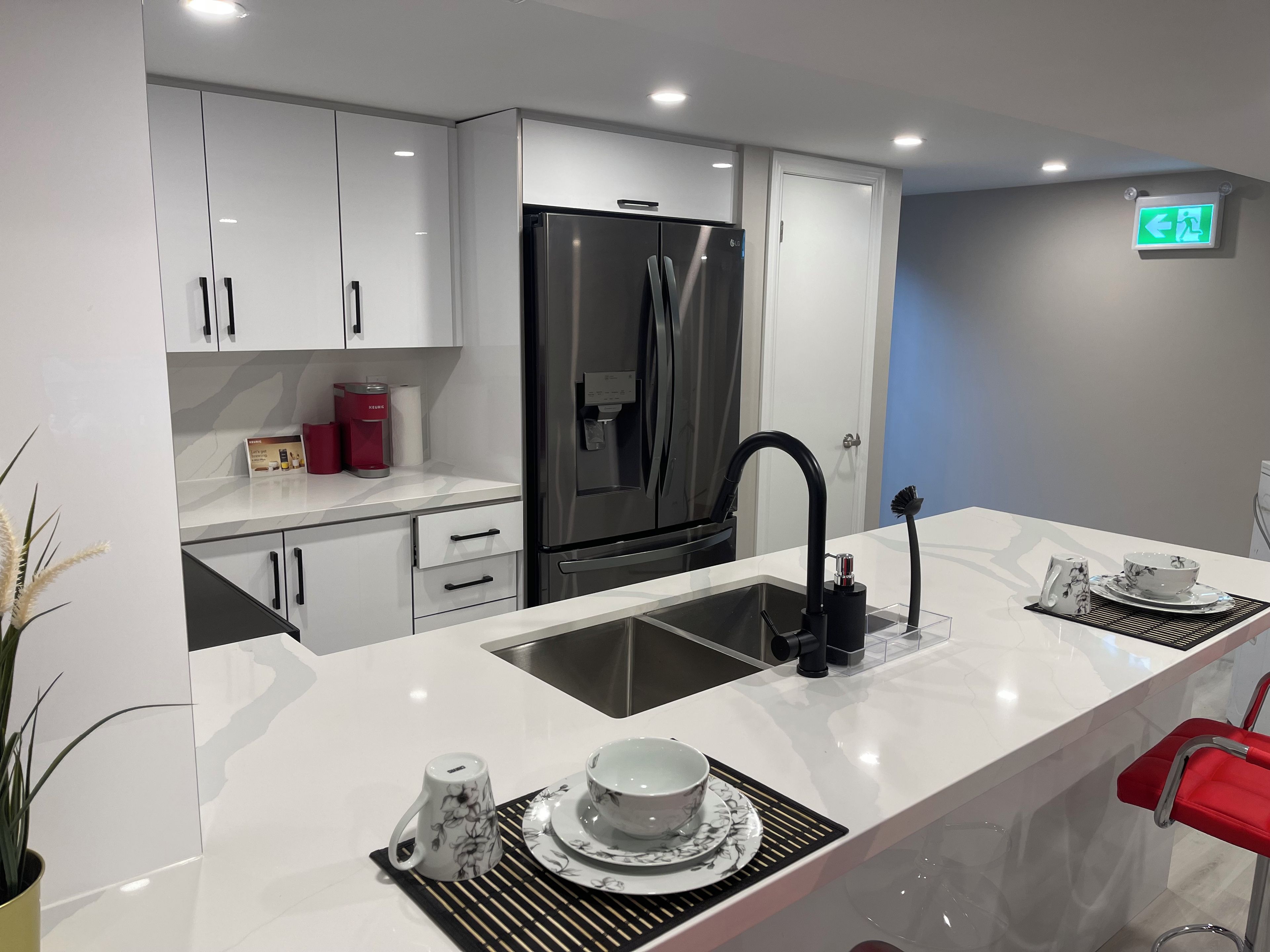 A modern kitchen with white cabinetry, a stainless steel refrigerator, a sink with a black faucet, and a dining area with a red chair.