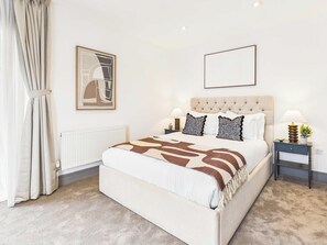 A bedroom with a large bed, a radiator, a nightstand with a lamp, and a framed picture on the wall. - Bright Westminster Duplex Apartment with Balcony (Greater London)