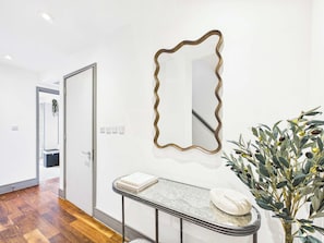 A hallway with a wooden floor, a door, a wall-mounted mirror, a small table with a book and a decorative item, and a potted plant with green leaves and olives. - Bright Westminster Duplex Apartment with Balcony (Greater London)