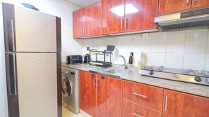 A compact kitchen with a stainless steel refrigerator, a washing machine, a gas stove, and wooden cabinets. - Boho Studio - Balcony & Sea View - Pool & Gym (Ajman)