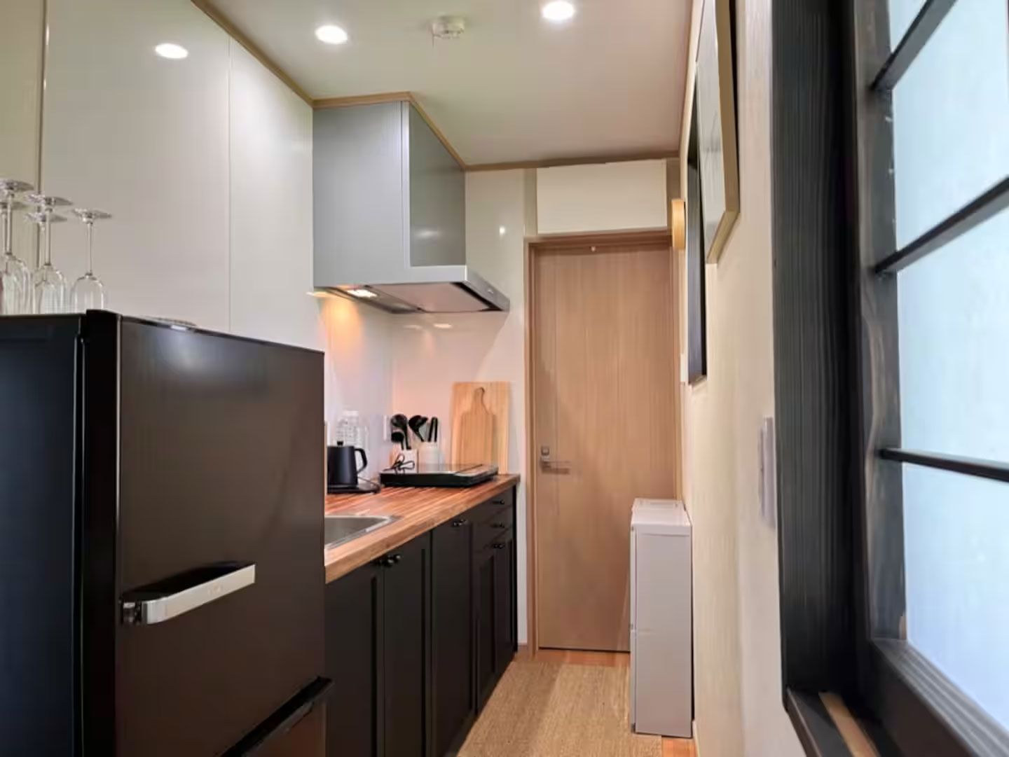 A compact kitchen with a refrigerator, a microwave, and a small oven. There is a wooden countertop with a sink and a window to the right.