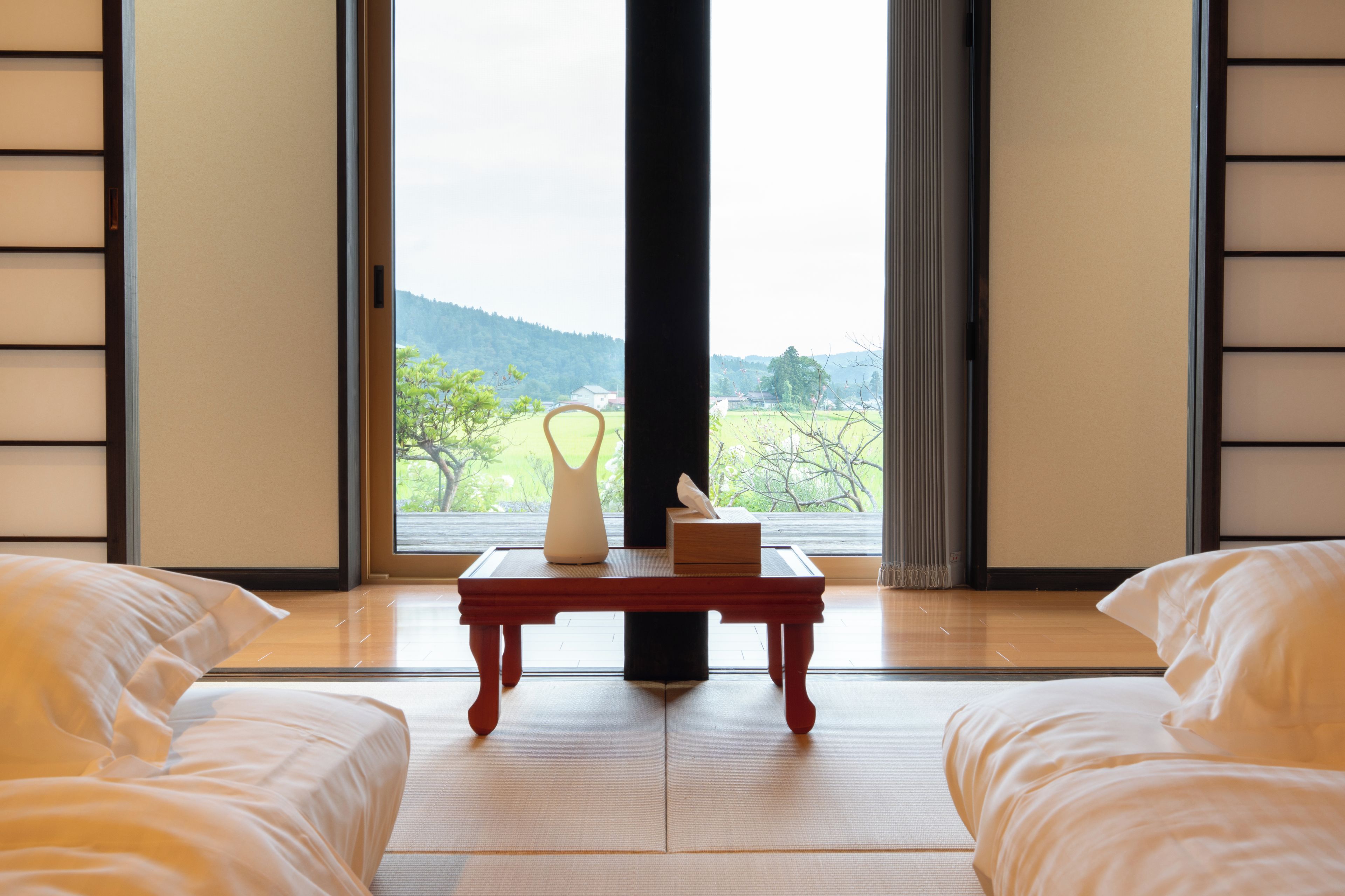 A room with a bed, a table with a vase and a box, and a view of a landscape through a window.