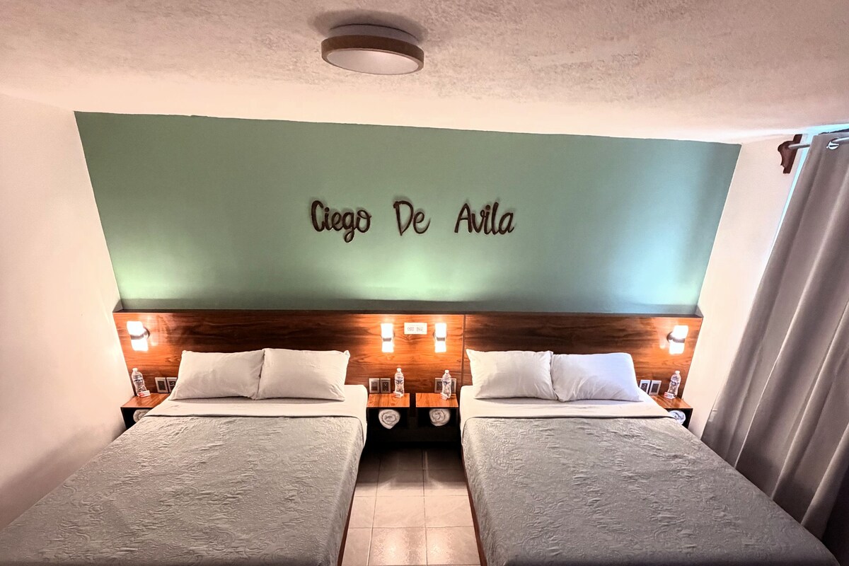 Habitación de hotel con dos camas, pared verde con el nombre 'Ciego De Avila' y una lámpara en el techo.