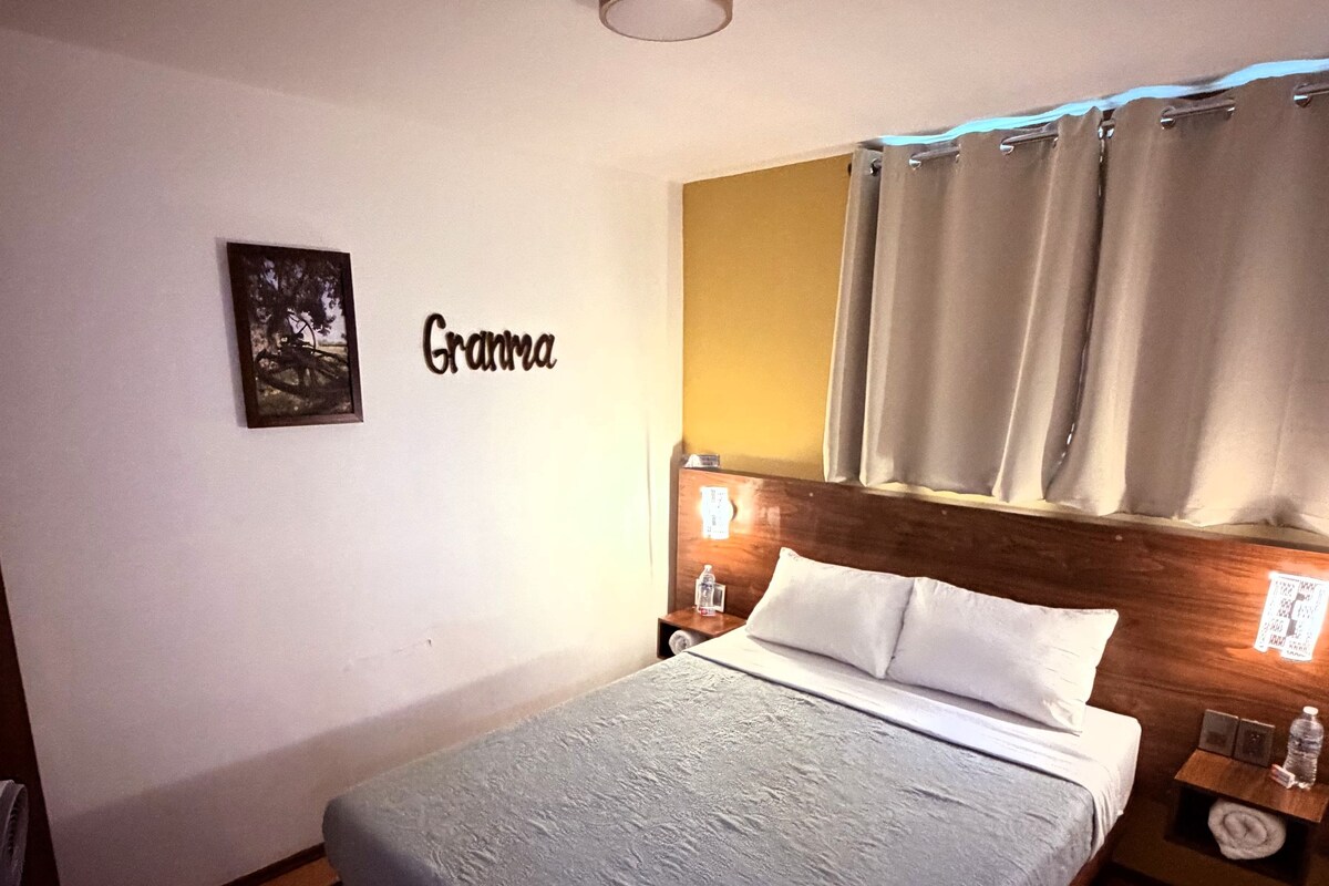 Un dormitorio con cama, cabecera, mesita de noche, lámpara de pared, un cuadro enmarcado y un cartel en la pared.