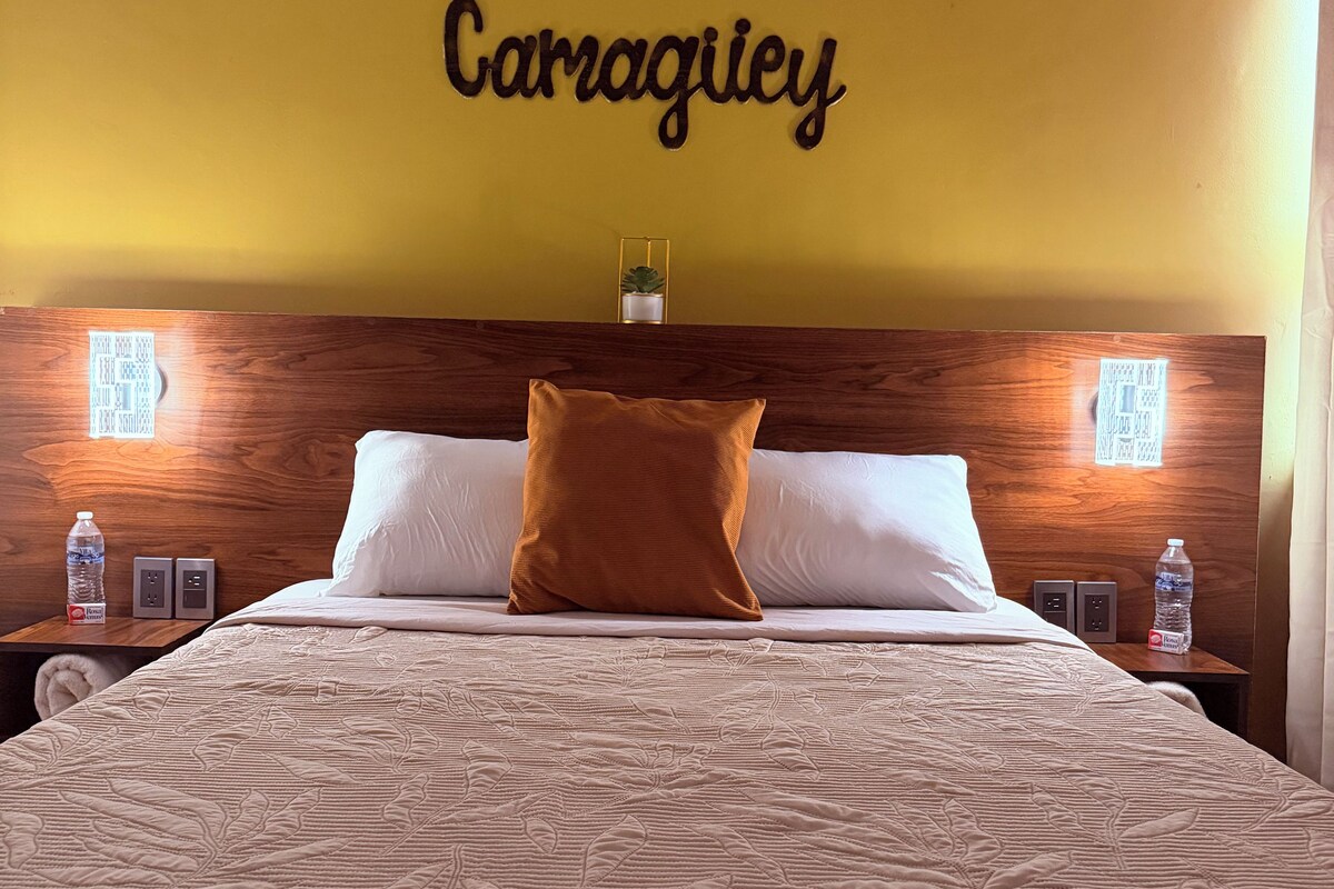 Habitación de hotel con cama, cabecera con placa con nombre, lámparas de noche y mesita de noche con botella de agua.