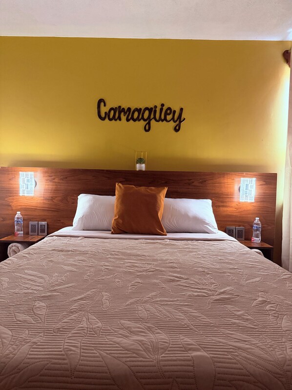 Habitación de hotel con cama, cabecera con placa con nombre, lámparas de noche y mesita de noche con botella de agua.